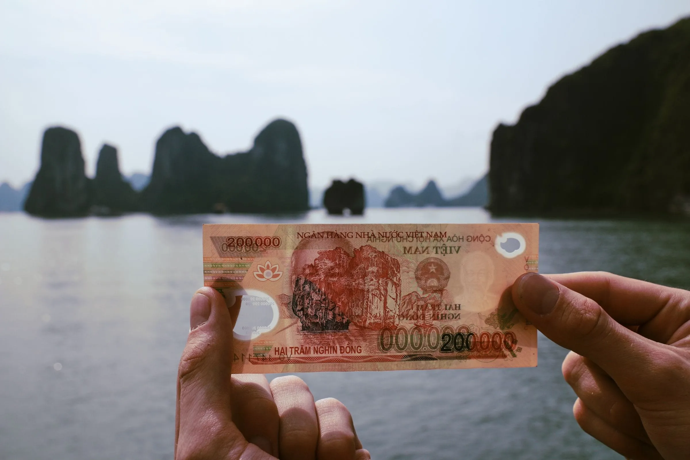 Hands holding a Vietnamese dong banknote in front of Ha Long Bay